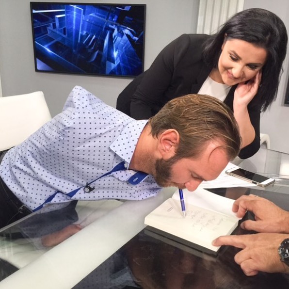 Nick Vujicic using his mouth to hold a pen and sign a book at Banja Luka while a smiling woman looks on