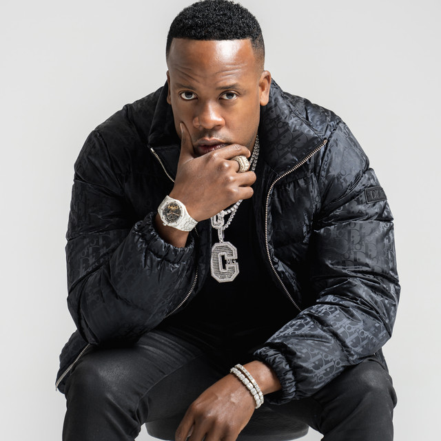 Yo Gotti wearing silver necklace with large letter C as pendant and other jewelries dressed all in black with his chin resting on his right hand