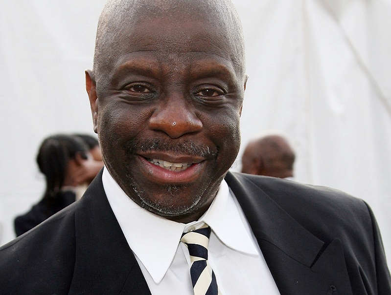 Jimmie Walker wearing a black coat with white shirt and striped tie