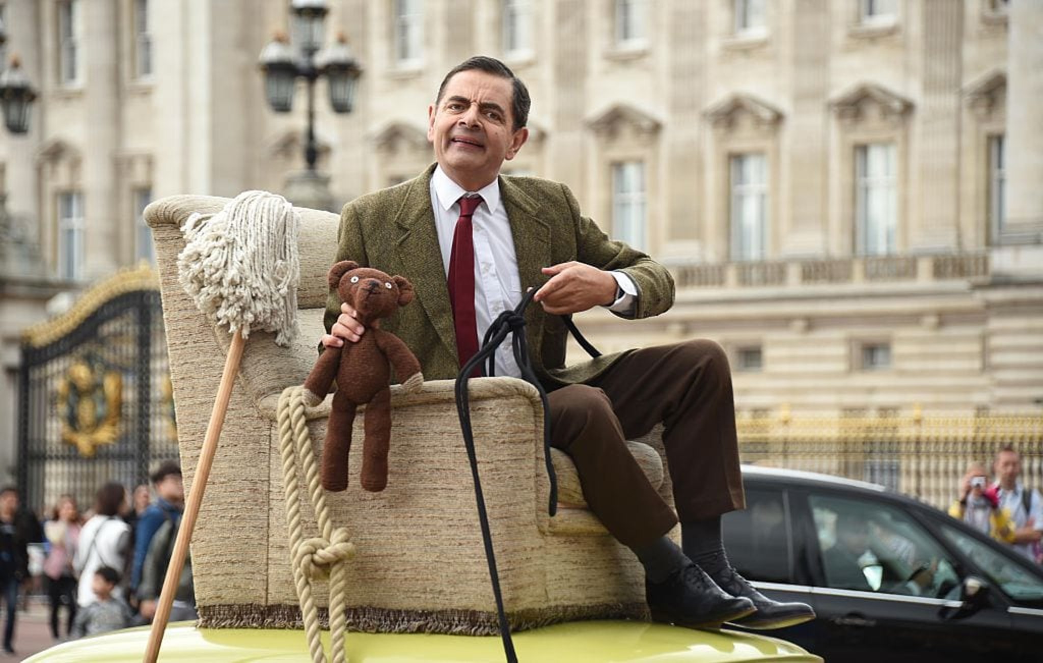 Rowan Atkinson portraying his role as Mr. Bean sitting on a chair on top of a car while holding Teddy