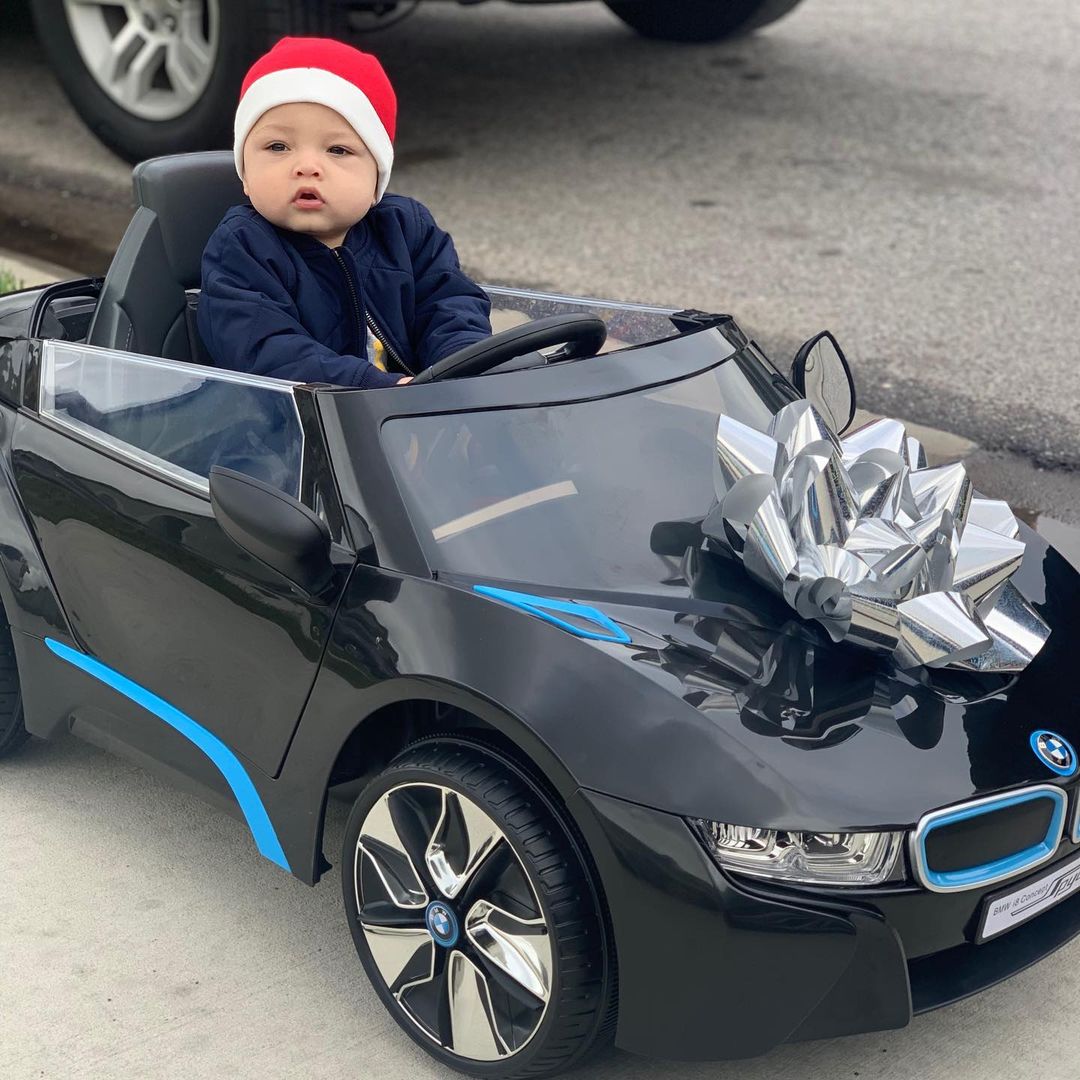Gekyume Onfroy in red Santa hat sits on Dynacraft BMW i8 concept ride-on toy car with giant silver foil ribbon on its hood