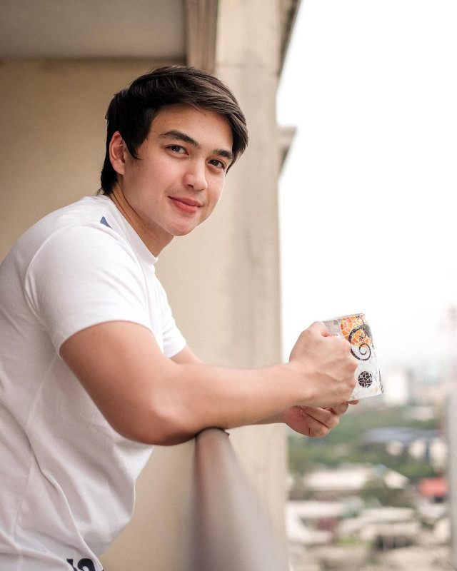 Dominic roque wearing a white shirt while holding a coffee mug in a balcony