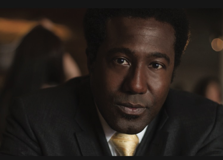 E Roger Mitchell in serious looks while wearing black suit and gold tie 