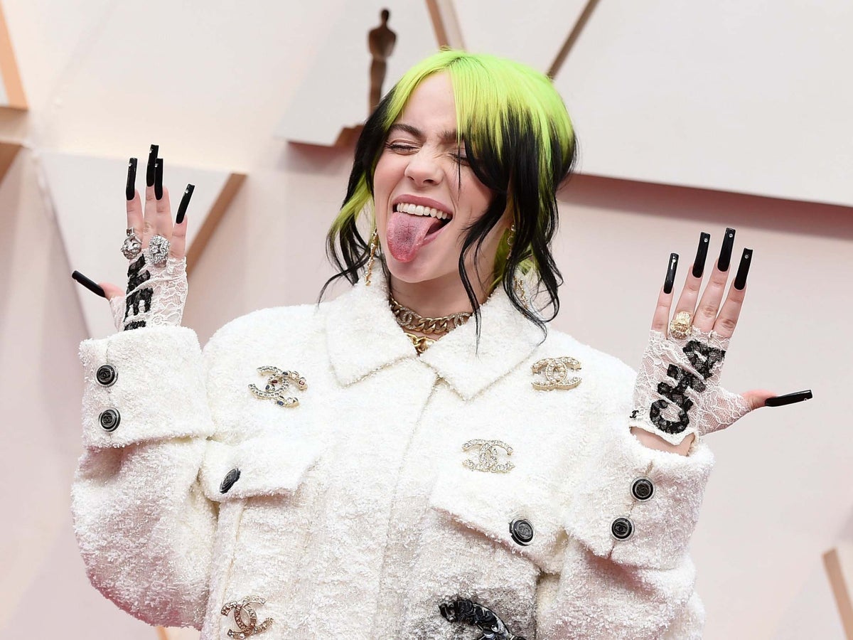 Billie Eilish wearing a white chanel jacket while sticking her tongue out and showing her black nails