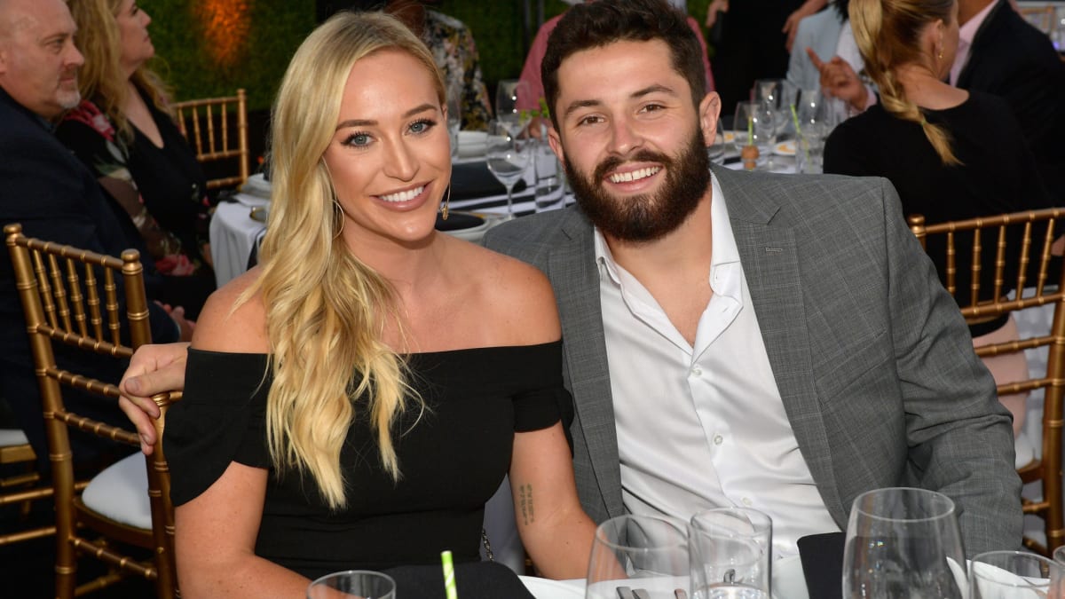 Baker Mayfield, Wearing A Grey Coat And White Shirt, Is Sitting With His Wife, Wearing A Black Dress.