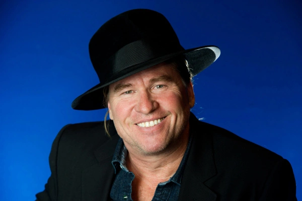 Val Kilmer wearing a dark jacket and hat while smiling