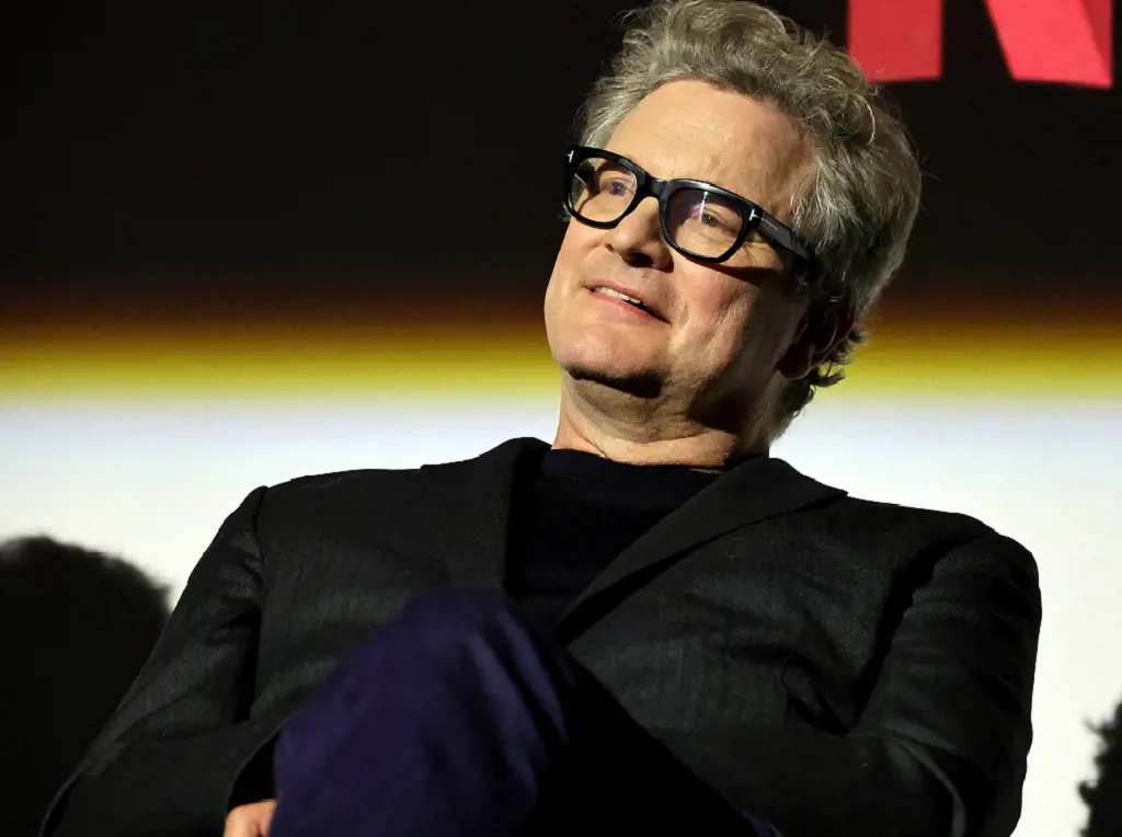 Colin Firth sitting down and wearing dark suit and large-rimmed glasses