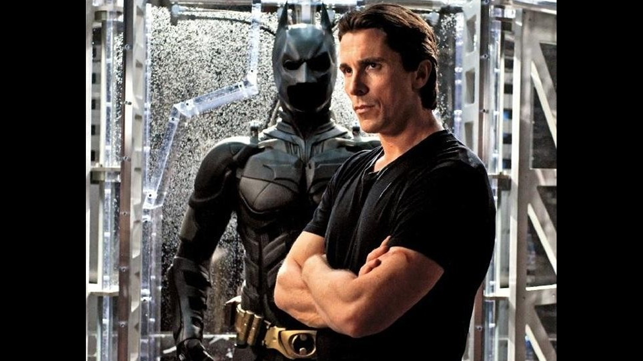Christian Bale in a black shirt standing infront of batman costume with crossed arms