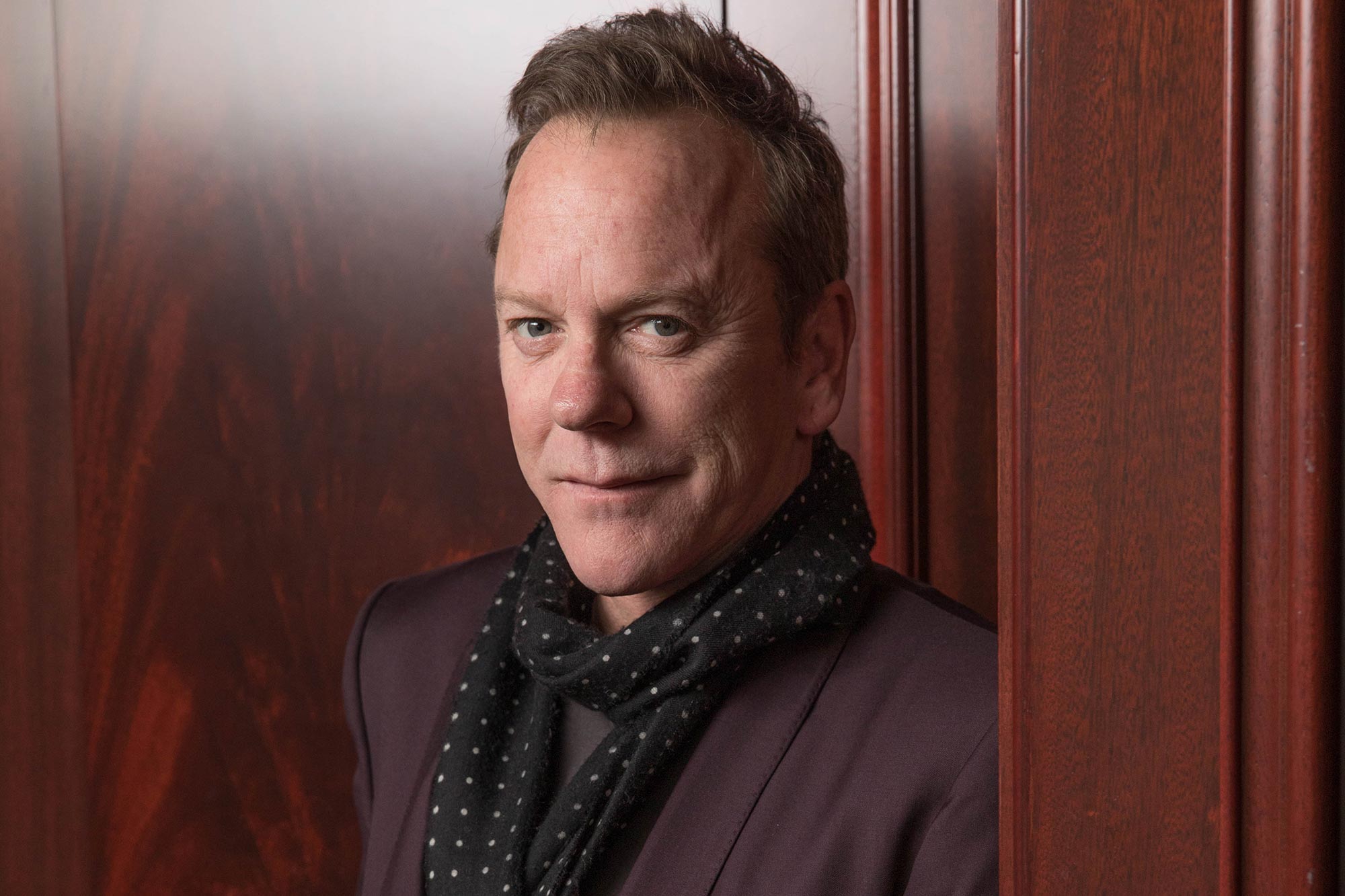 Kiefer Sutherland wearing a dark glossy suit and black scarf while smiling