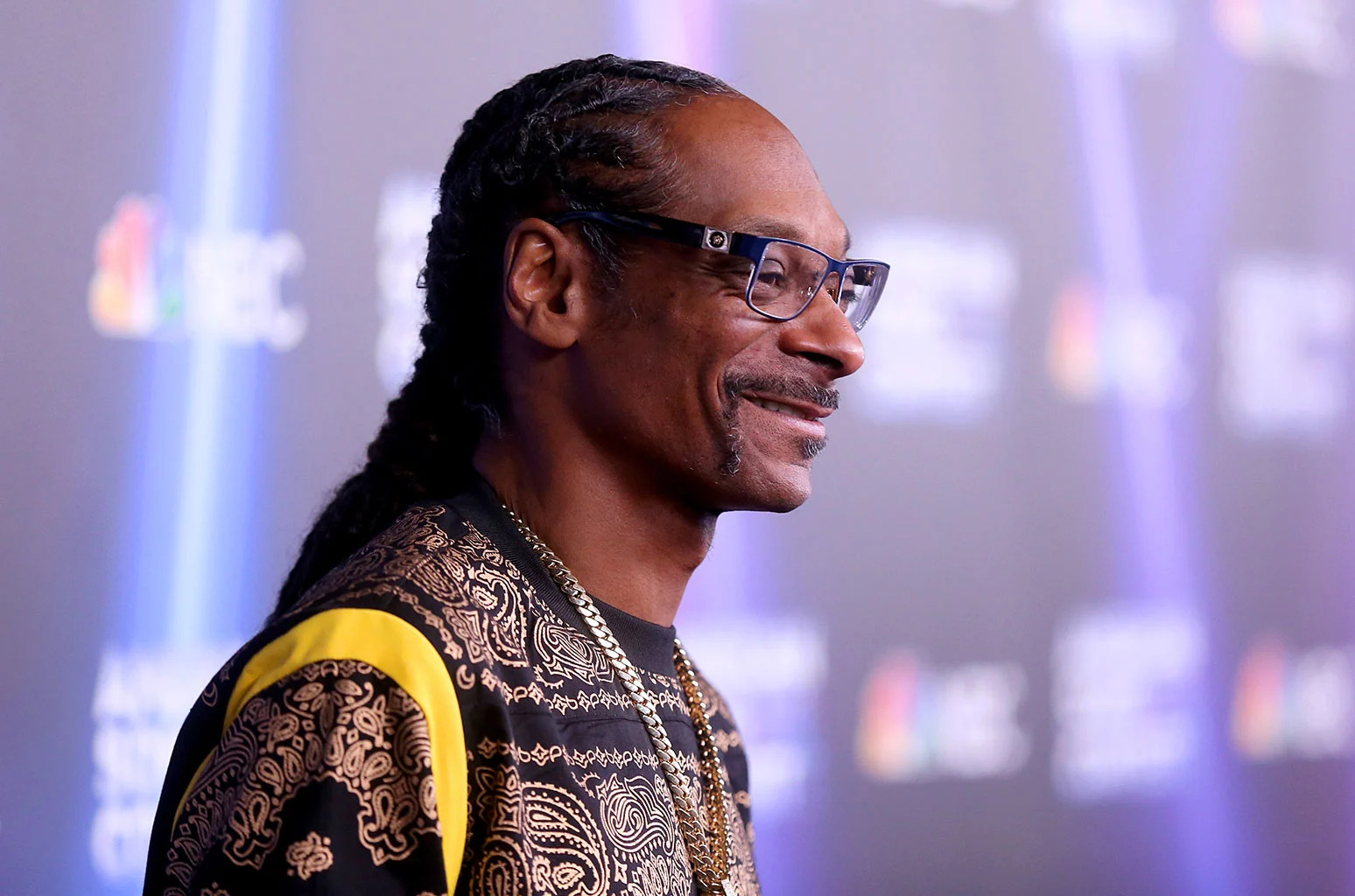 Snoop Dogg wearing a black printed shirt, gold chain necklace, and eyeglass