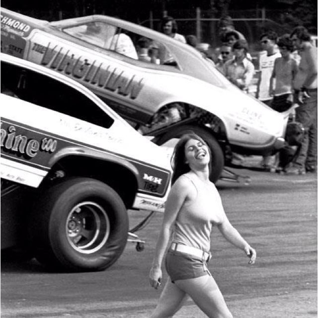 Pam hardy is looking very happy and joyful as always on a racing car field with race cars and fans behind her