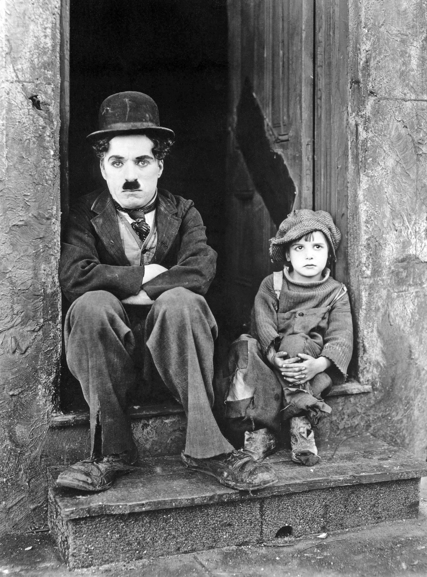 Charlie Chaplin sitting with crossed arms at a small entrance with a child