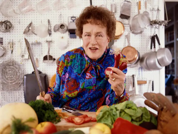 Julia Child holding a red onion and long knife with more cooking tools on the wall behind her