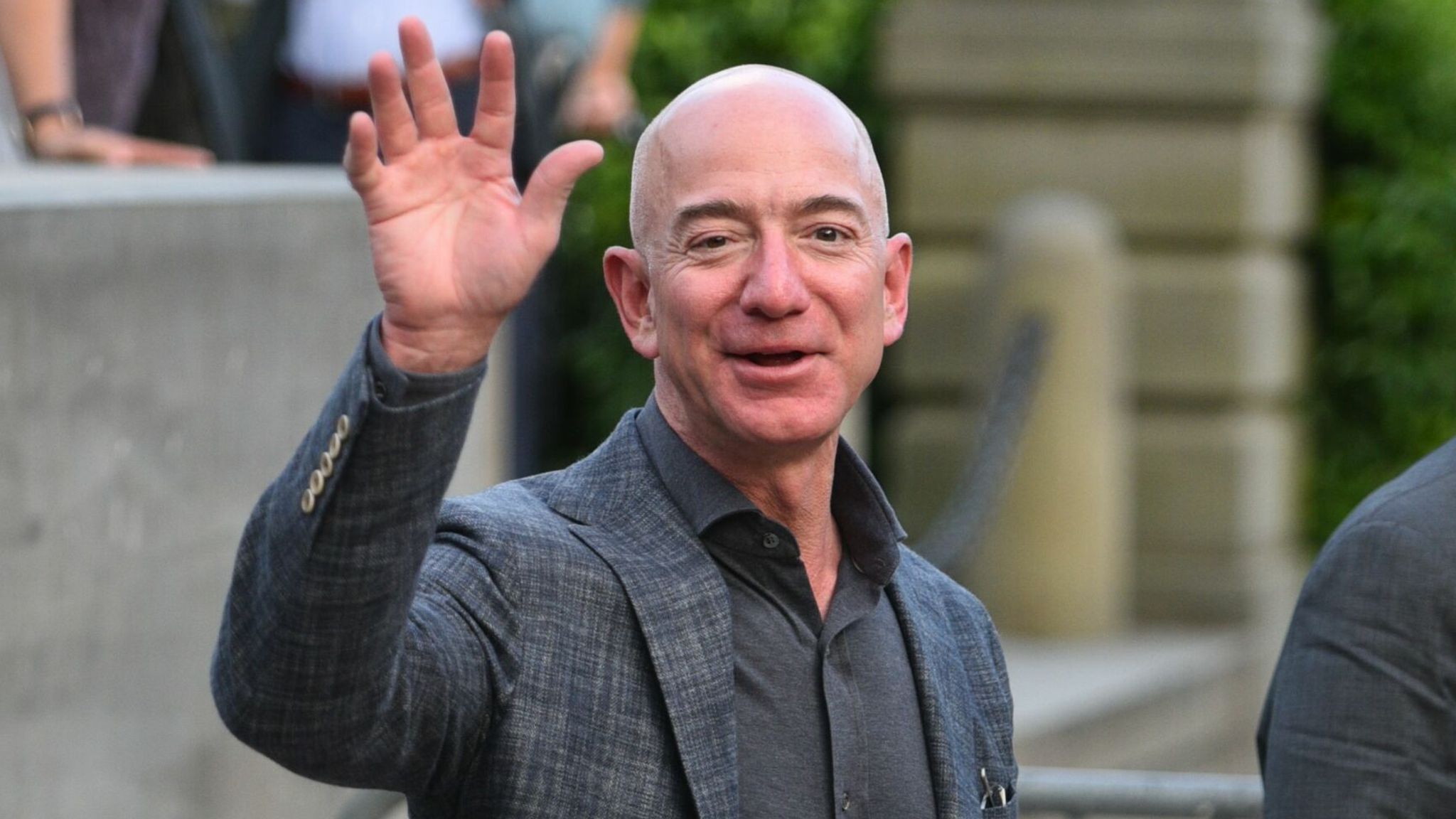 Jeff Bezos waving his hand whil wearing a gray suit