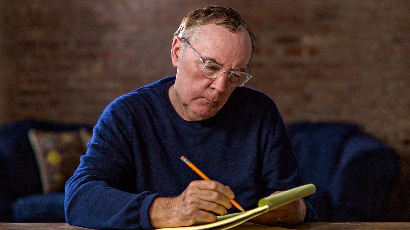 James Patterson wearing a blue sweater while writing on paper