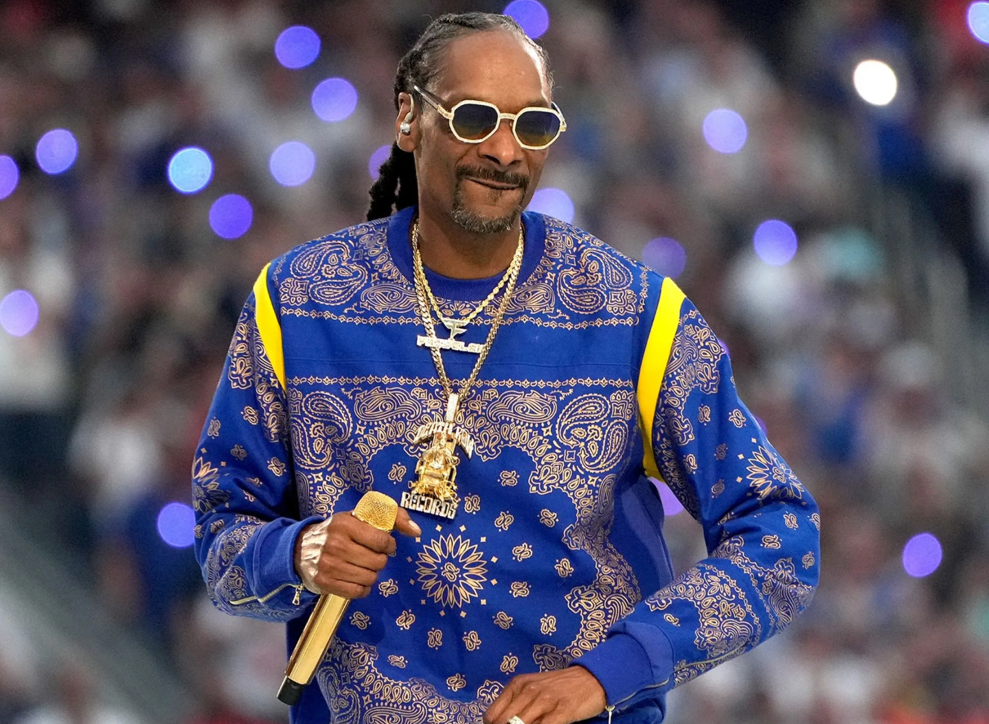 Snoop Dogg wearing a blue bandana patterned sweater while holding a gold microphone