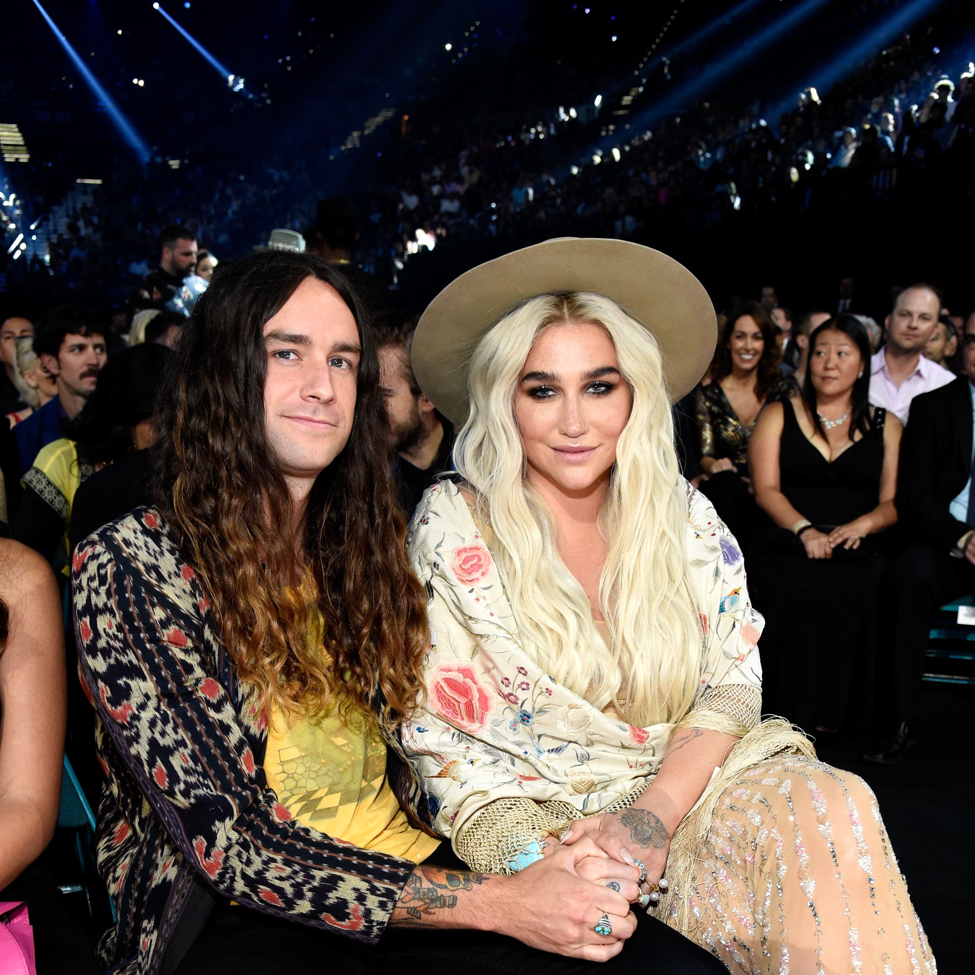 Brad Ashenfelter wearing a yellow shirt and patterned sweater and Kesha wearing a floral scarrf and nude dress
