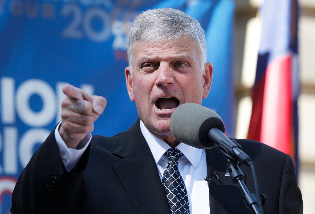 Franklin Graham wearing a black suit on a white shirt and blue tie as he speaks into a microphone while pointing