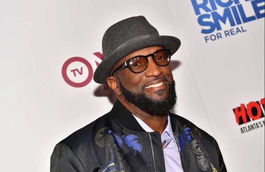 Rickey Smiley wearing a black jacket, a hat, and a black eye glass at an event