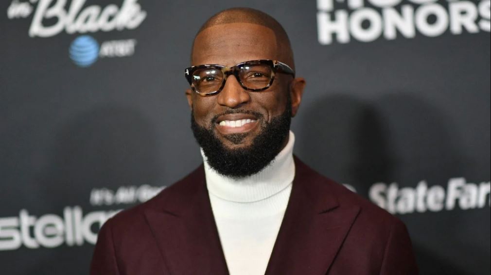 Rickey Smiley wearing a brown suit and an eye glass at an event