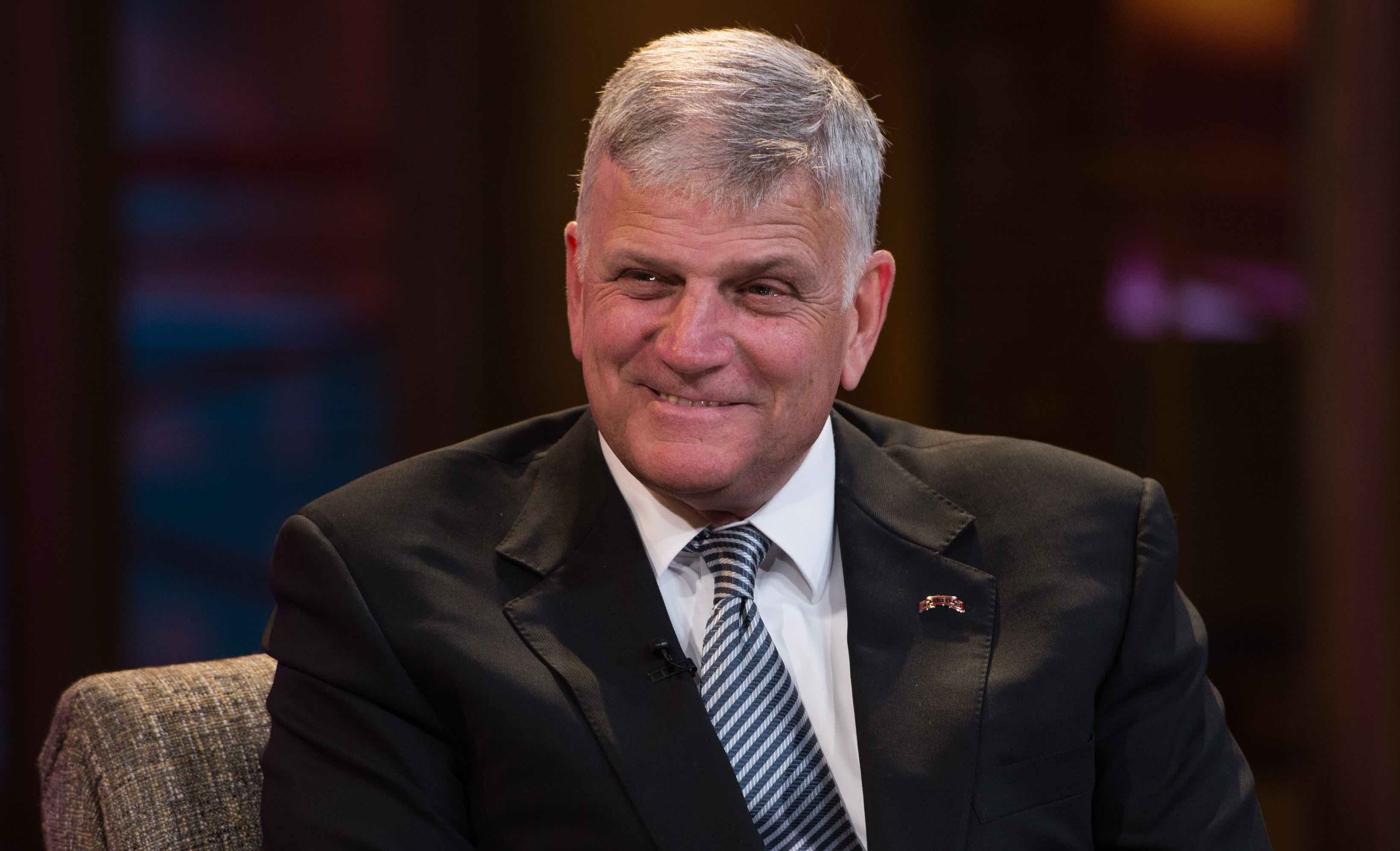 Franklin Graham wearing a black suit on a white shirt and a blue tie with a smile on his face