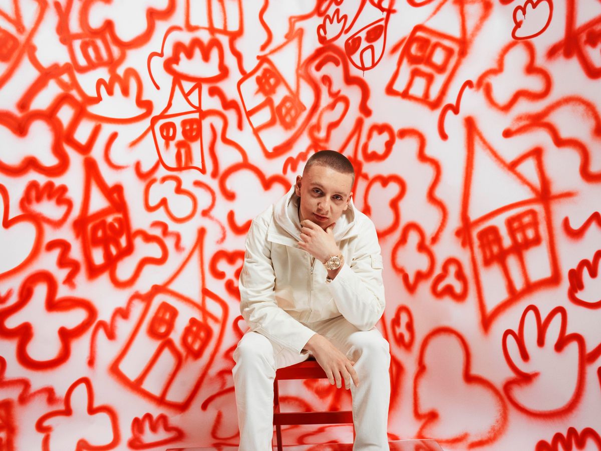 Aitch wearing a white jacket and pants with red drawings on the wall