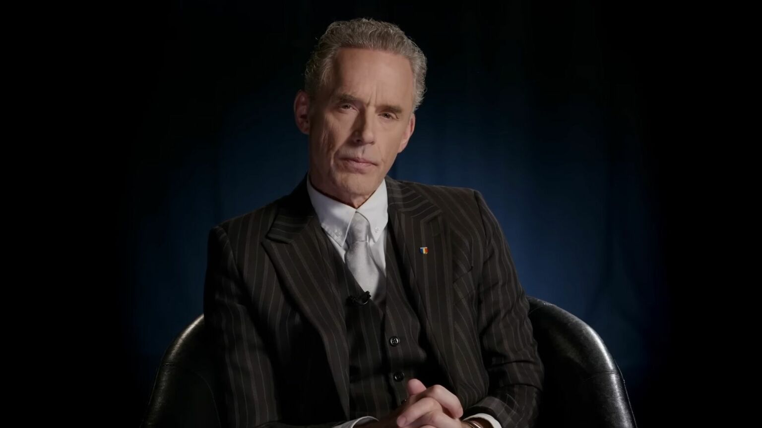 Jordan Peterson wearing a suit on a white shirt sitting on a chair