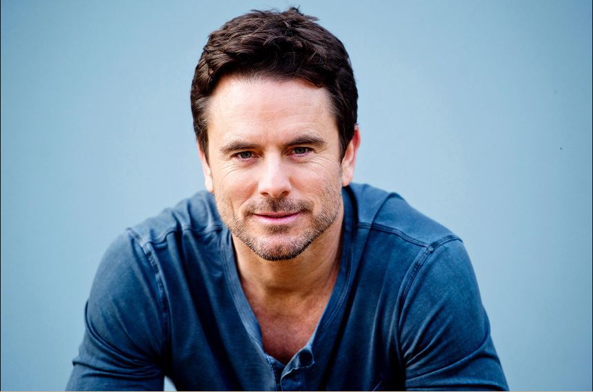 Charles Esten wearing a blue t-shirt with a little smile on his face