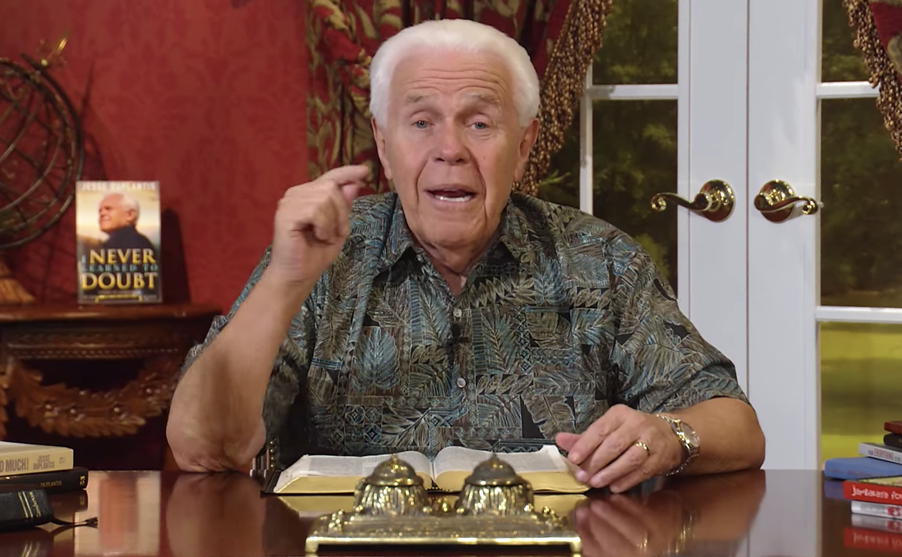 Jesse Duplantis sitting at a table with a Bible as he preaches