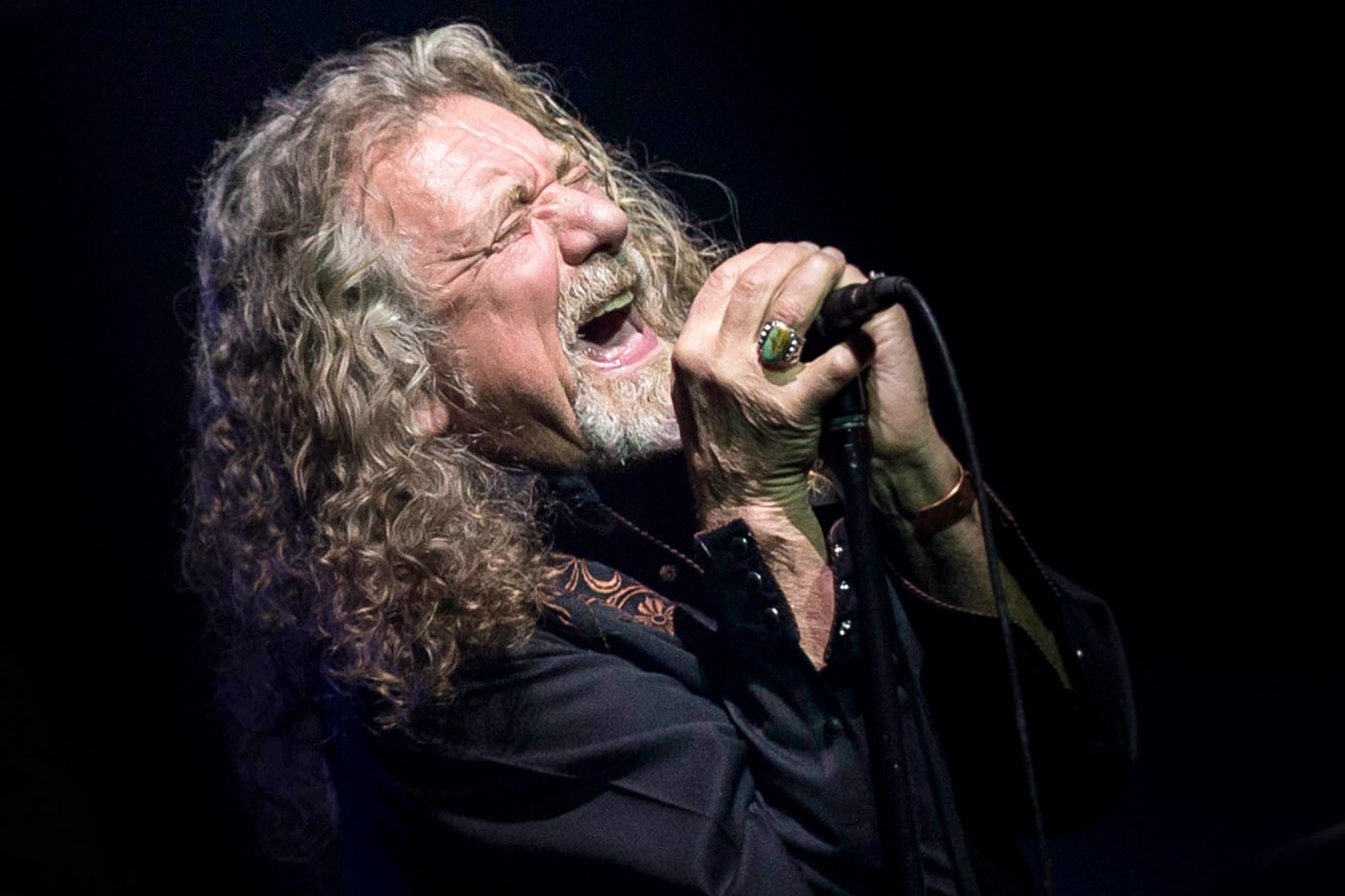 Robert Plant holding a mic whilee wearing a black polo long sleeves