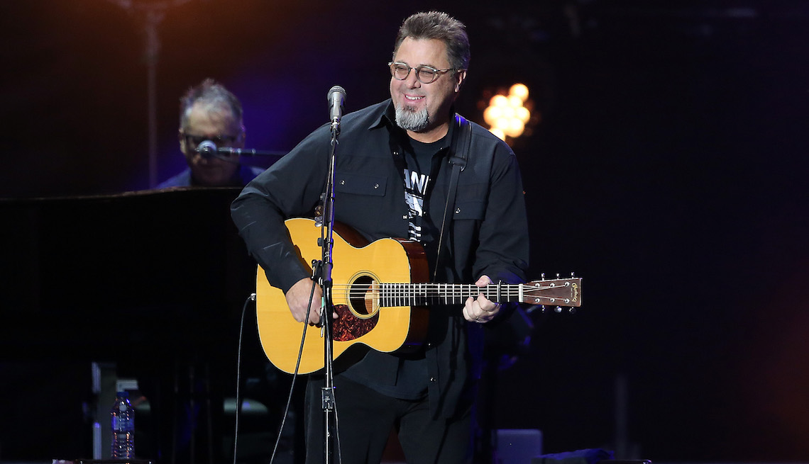Vince Gill plays a guitar as he sings into a microphone