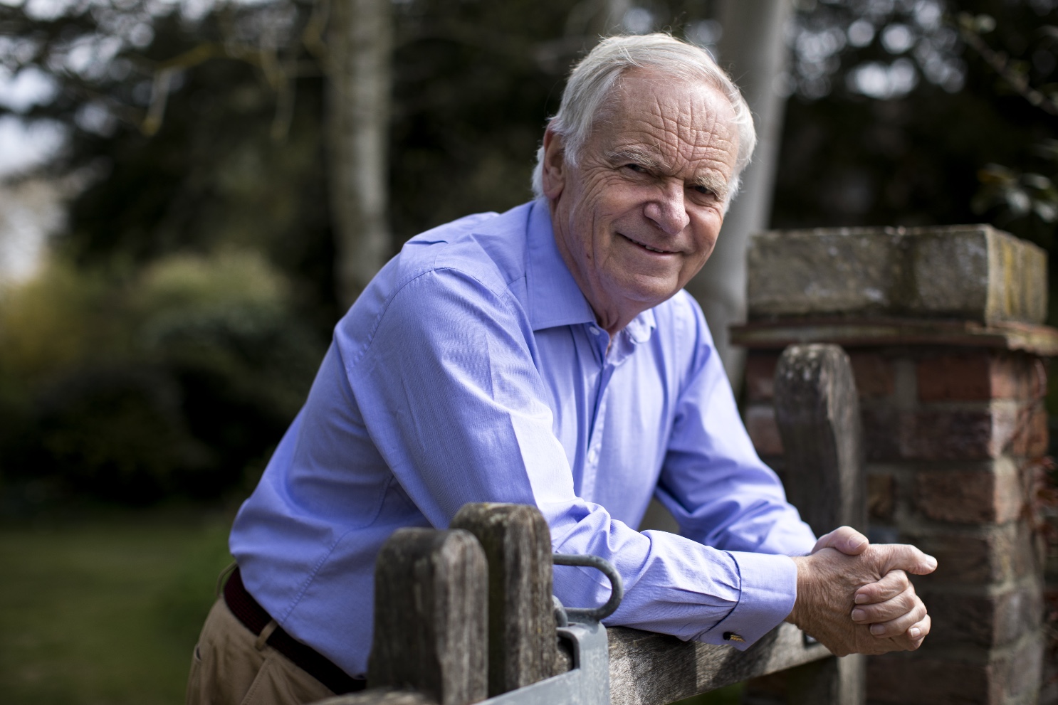 Jeffrey Archer wearing a light blue polo long sleeves while leaning on a wooden gate
