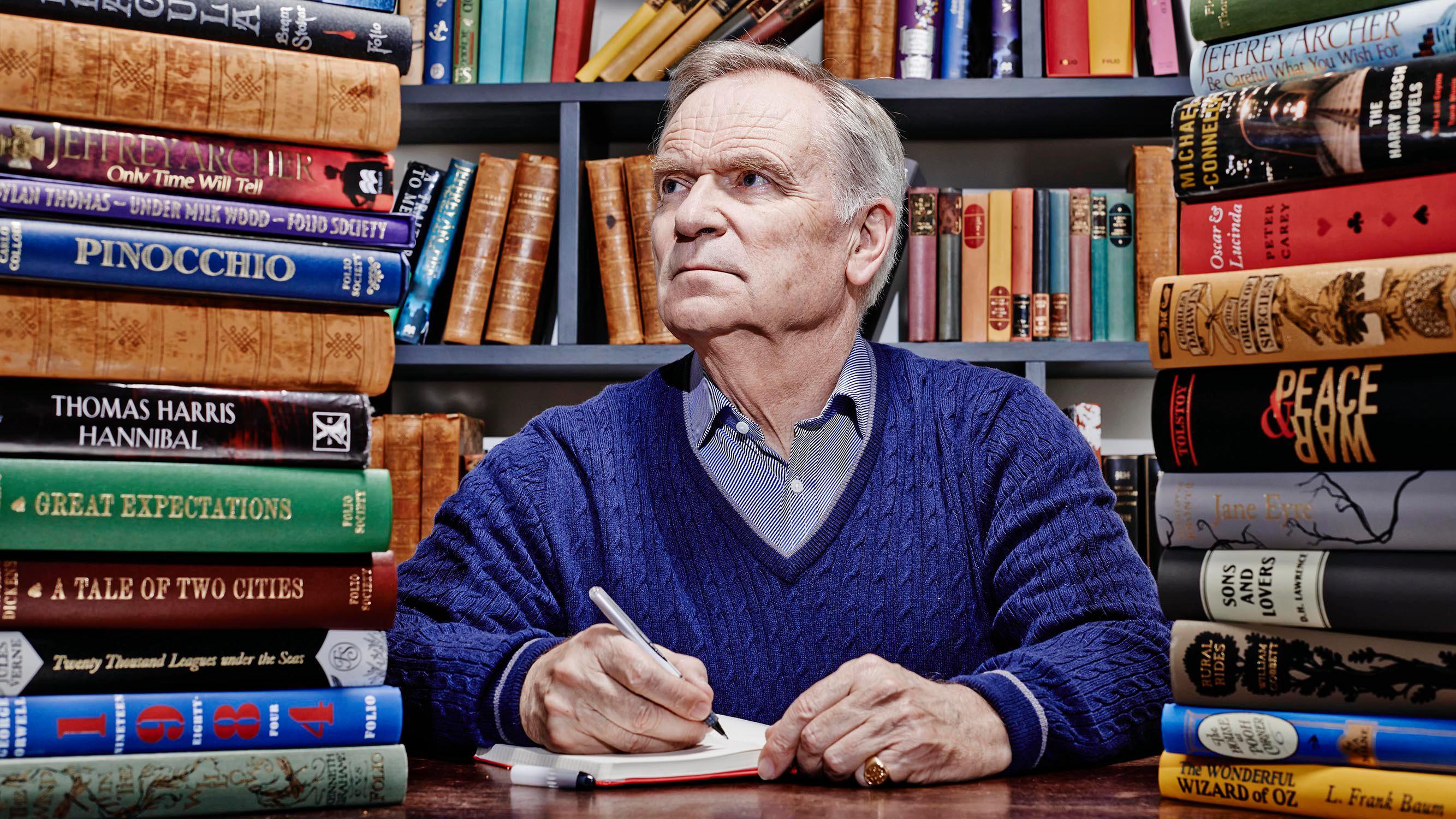 Jeffrey Archer wearing a blue sweater while holding a pen and notebook
