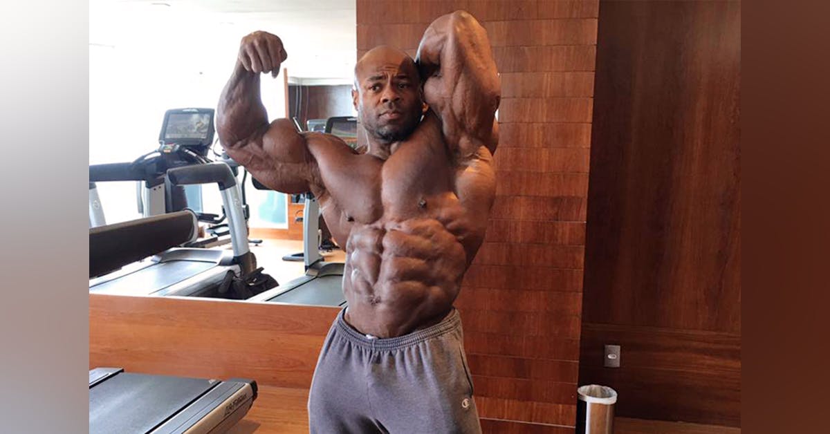 Kai Greene flexing his muscles in the mirror while wearing a gray sweatshorts