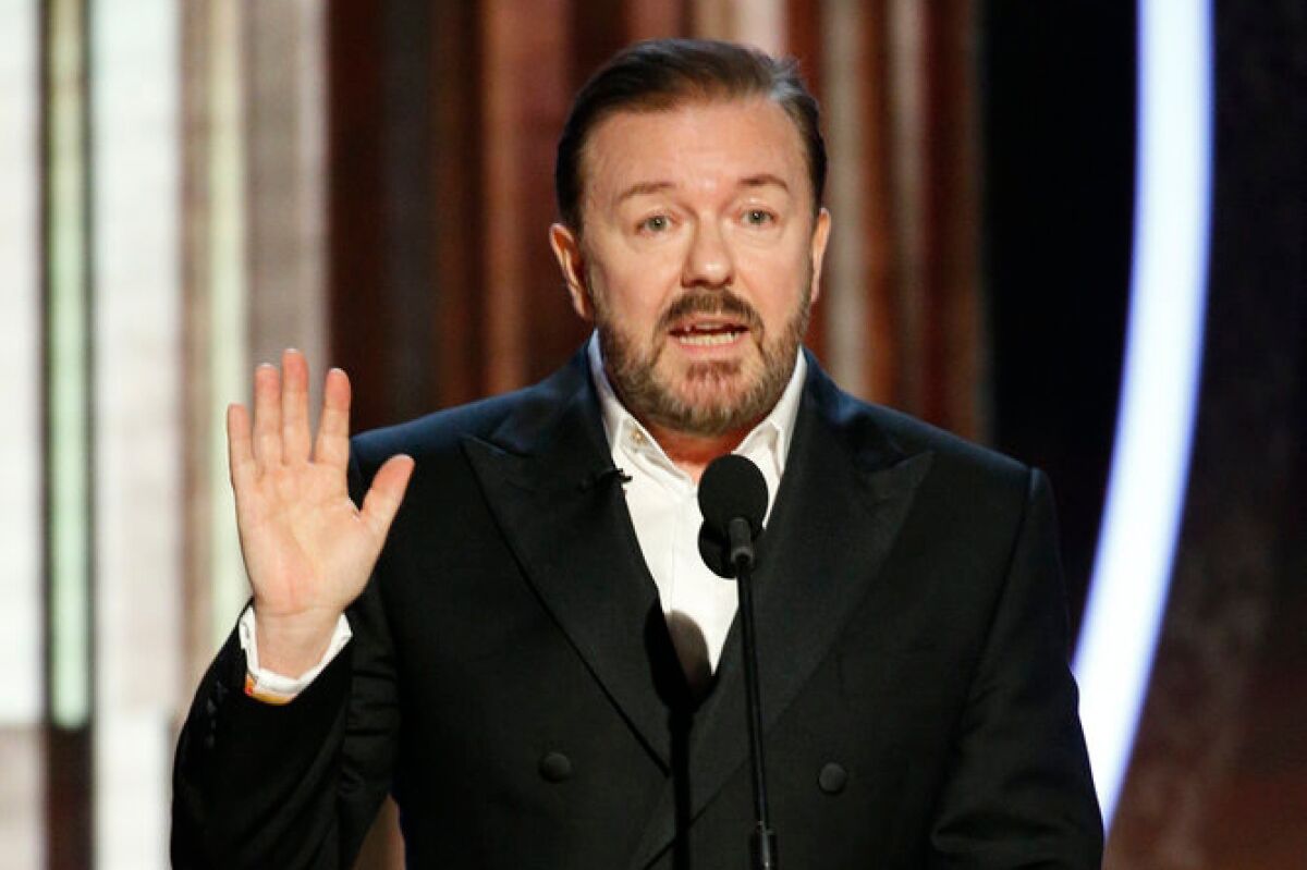Ricky Gervais wearing a black suit while raising his hand
