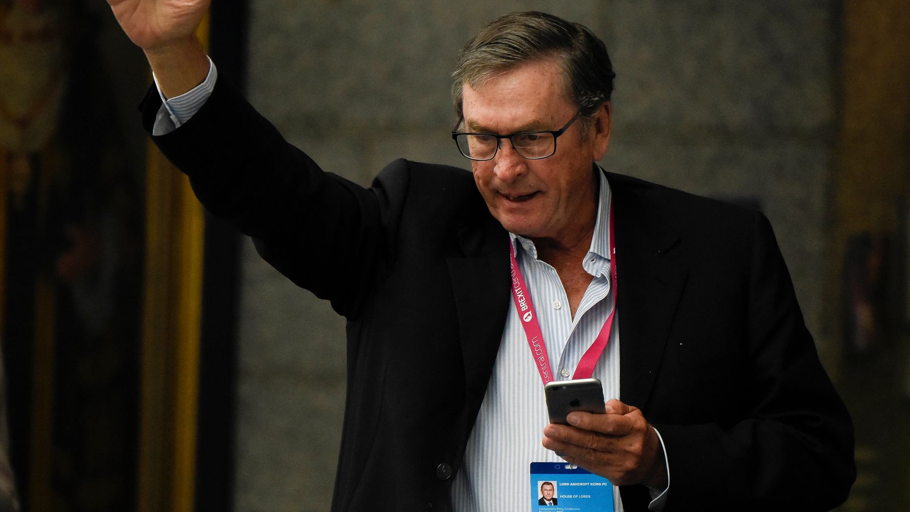 Michael Ashcroft wearing a black coat while raising his hand and holding a phone