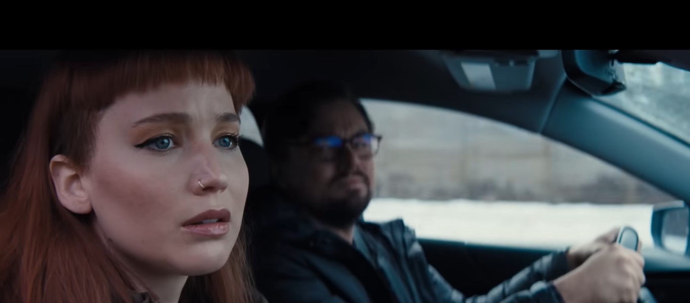 Jennifer Lawrence with a fake nose ring inside a car beside a bespectacled Leonardo DiCaprio who is driving