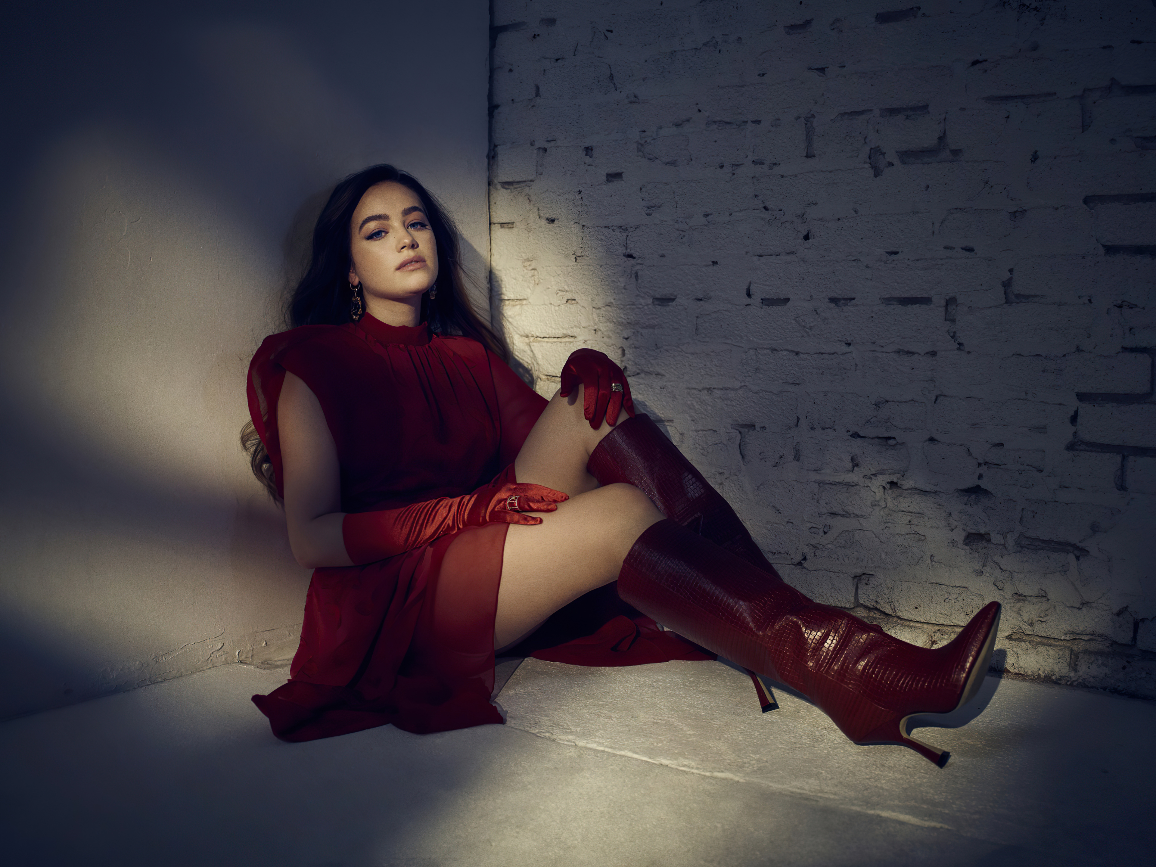 Mary Mouser wearing a red dress, gloves, and boots while sitting