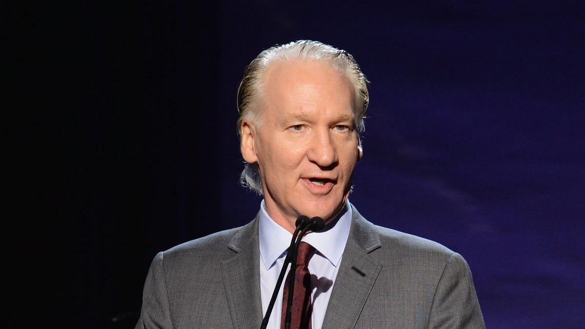 Bill Maher wearing a gray suit while talking on a mic