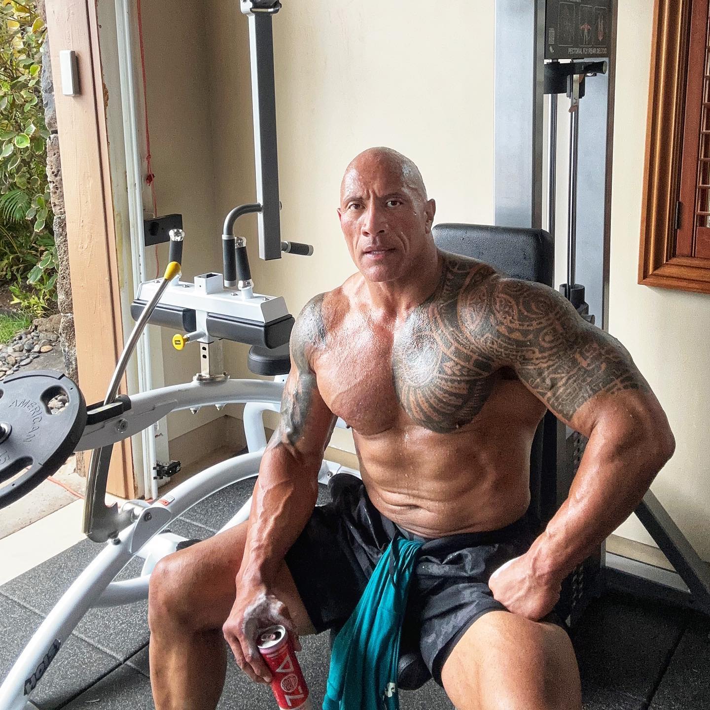 A shirtless Dwayne Johnson in his home gym in black shorts holding a can of ZOA energy drink