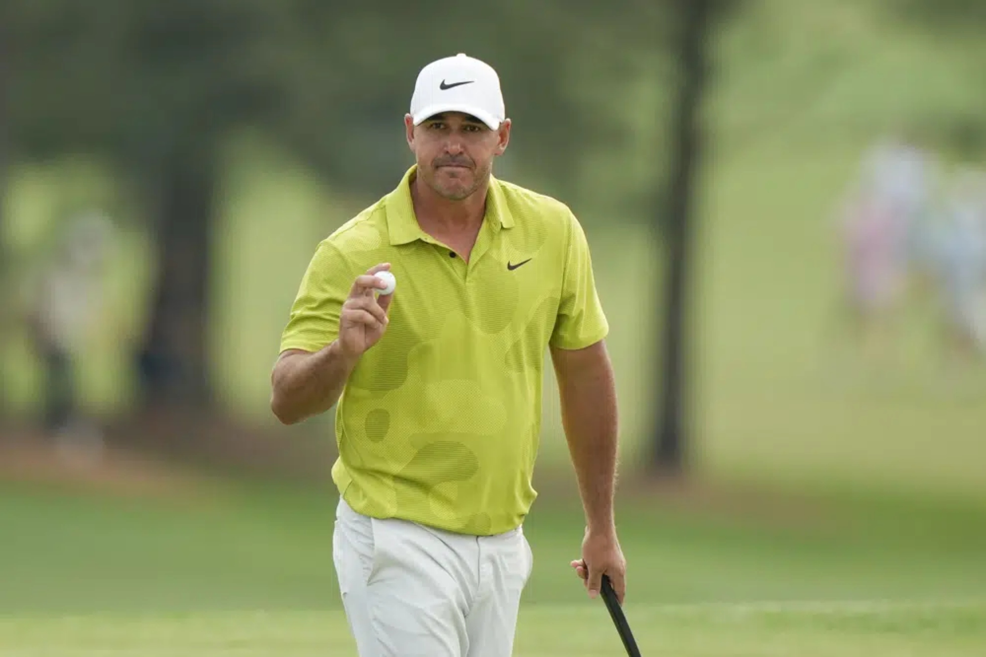 Brooks Koepka wearing green shirt and white cap while holding a golf ball