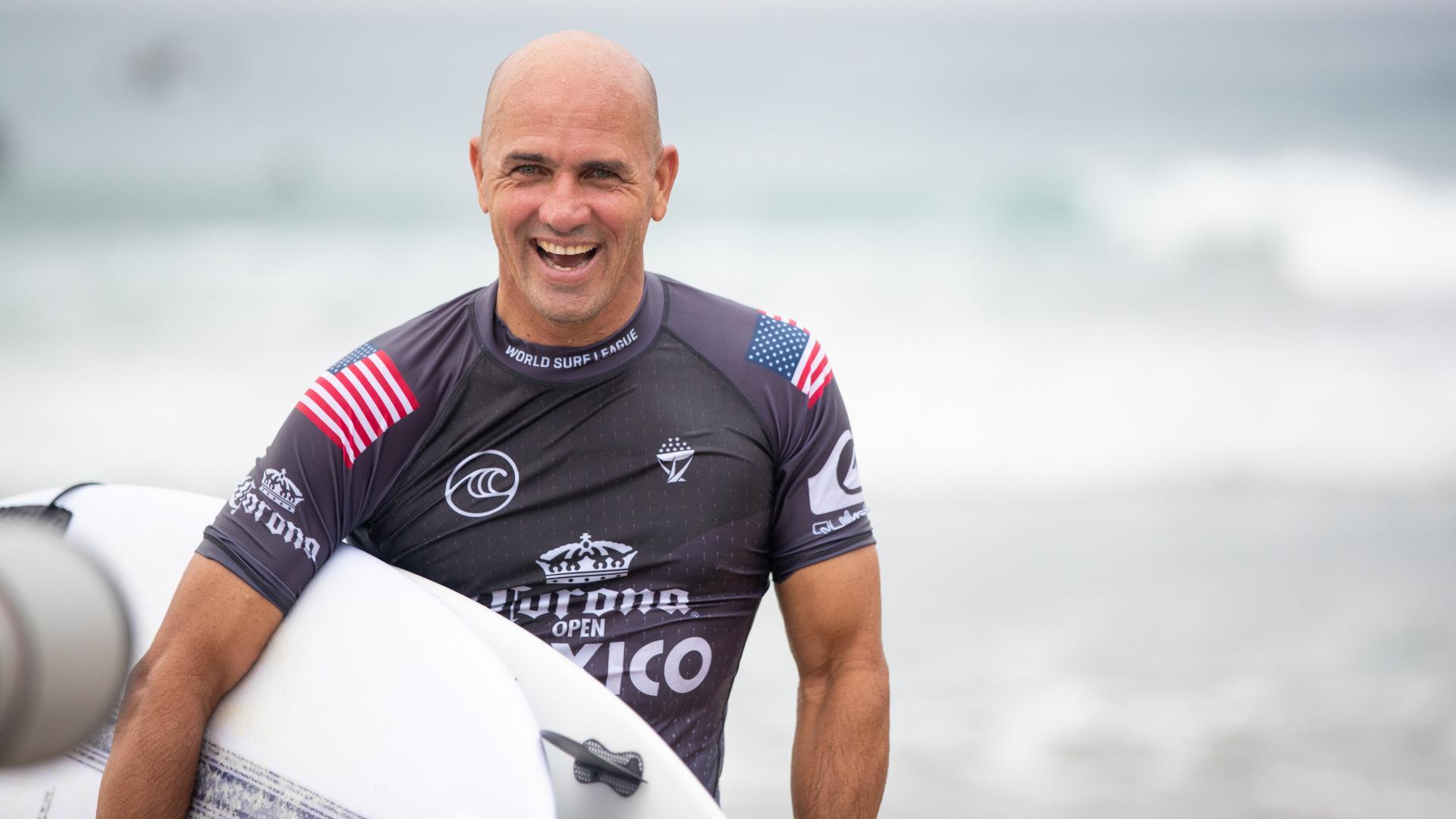 Kelly Slater wearing a black shirt hile holding a white surfboard