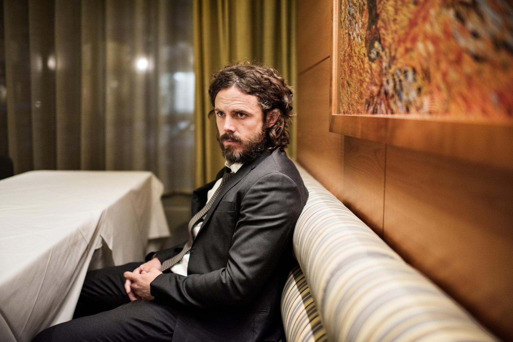 Casey Affleck sitting on a sofa wearing a black suit and pants