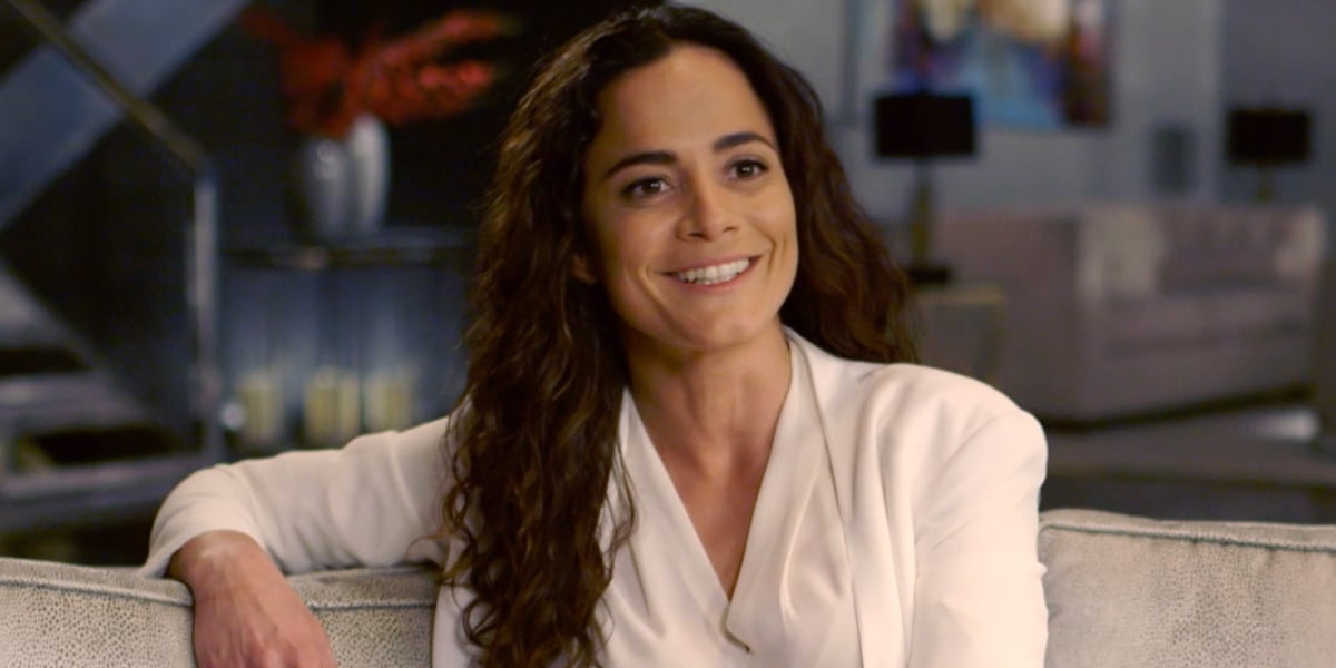 Alice Braga wearing a white gown while sitting on a couch with a smile on her face